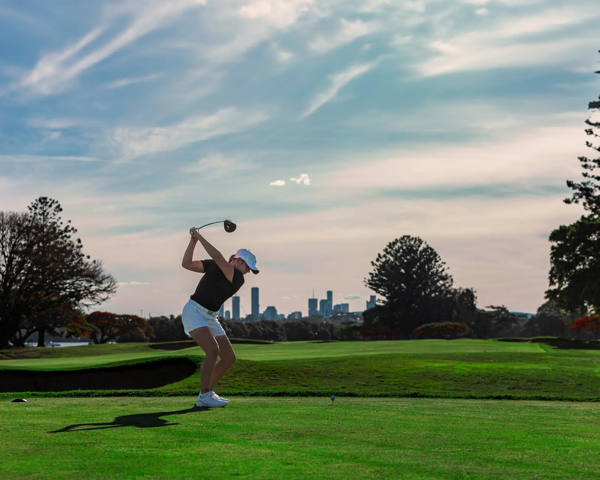 Female golfer in a black top and white shorts preparing to drive a golf ball on a course with a city skyline in the background, showcasing the GolfN app's focus on enhancing the golfing experience.
